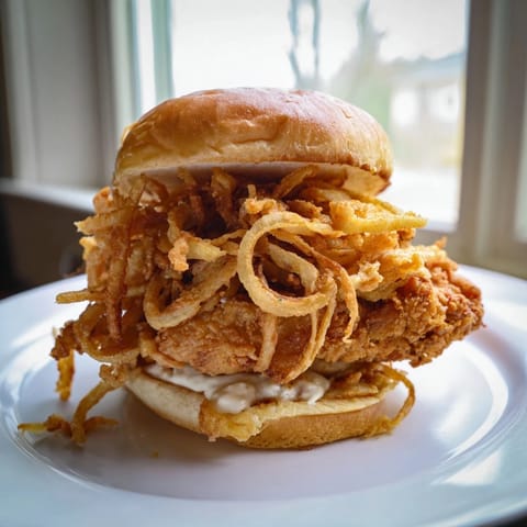 Homemade Crispy Onion Chicken Sandwich served with creamy aioli on a soft bun, featuring golden fried onions and tender chicken.  